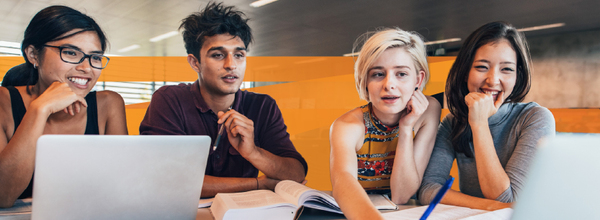 Higher education students at a large desk with computers and book.