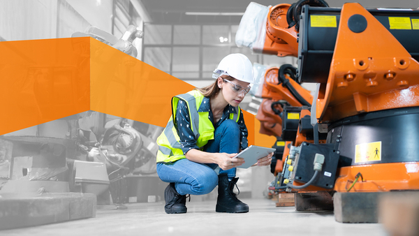 A woman wearing protective gear looking at a tablet, kneeling next to a row of heavy machinery